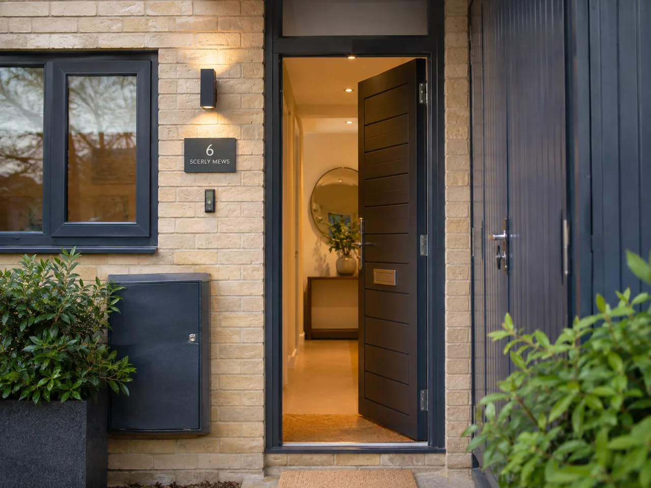 Open dark front door of a modern brick Oxford mews house, with warm light in the hallway beyond