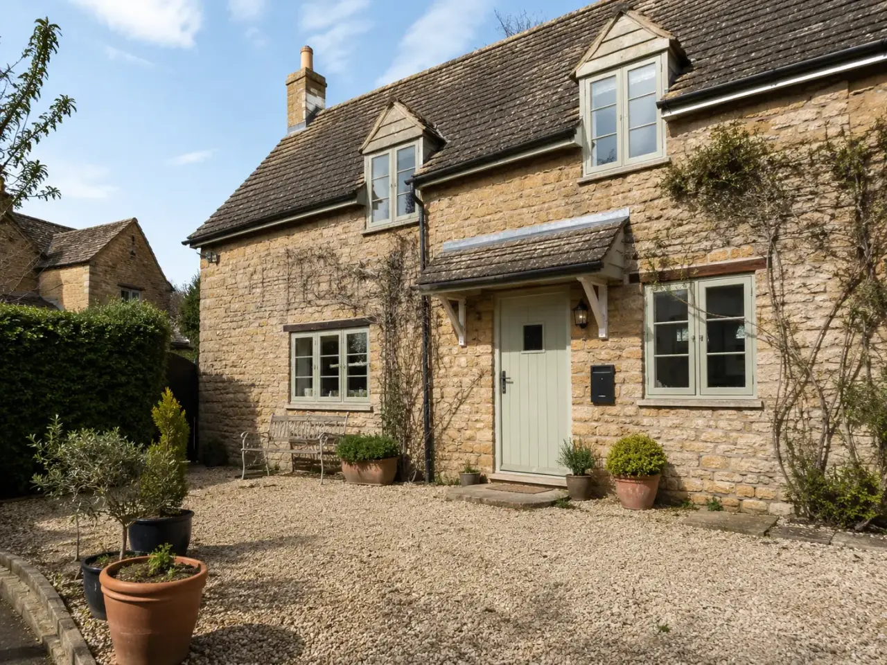 Cotswold stone cottage with dormer windows, a pale green front door and a gravel forecourt with potted plants
